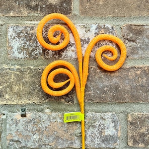 May include: An orange decorative floral stem with three spiraling curls. The stem is a bright, solid orange color, and the curls have a textured, glittery appearance. The stem is set against a weathered brick wall.