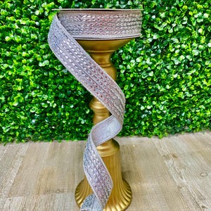 Op de afbeelding: Een gouden metalen decoratieve standaard met een ronde bovenkant en drie poten. Een zilveren lint met een parelmoerontwerp is om de standaard gewikkeld.