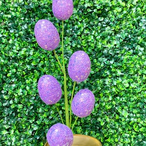 May include: A decorative Easter egg stem featuring six purple sequined eggs. The eggs are attached to green stems and sit in a gold-colored, decorative bowl. The background is a dense, green hedge.
