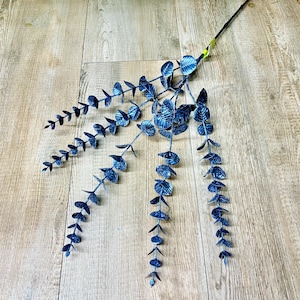 May include: A single stem of artificial eucalyptus in a deep blue hue. The leaves are small, round, and arranged along the stem in a cascading pattern. The stem is dark green and the leaves have a textured appearance. The eucalyptus is placed on a wooden surface.