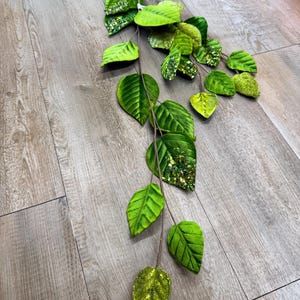 May include: An artificial leaf stem featuring green leaves in various shades, with some leaves having a glittery finish. The stem is brown and has a hook. The leaves are arranged in a cascading design, suitable for decoration.