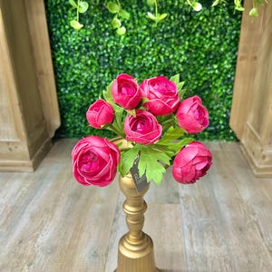 May include: A gold-coloured decorative candle holder with a flared base, holding a bouquet of vibrant pink artificial flowers. The flowers have green leaves and are set against a green backdrop.