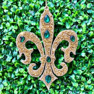 Puede incluir: Adorno de color dorado en forma de flor de lis con detalles de gemas verdes.