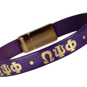 May include: A purple leather bracelet featuring the gold Greek letters Omega Psi Phi repeated along the band, separated by small gold dots. The bracelet is secured with a rectangular metallic gold magnetic clasp closure.