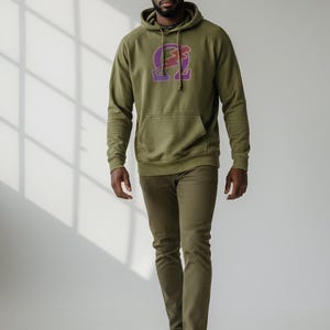 May include: Olive green hoodie with a kangaroo pocket and a purple and pink graphic on the front. The hoodie is paired with olive green pants and white sneakers. The person is walking towards the camera.