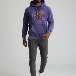May include: A purple hooded sweatshirt with a front pocket and a gold graphic design. The graphic is a stylized letter and lightning bolt. The person is wearing grey trousers and white trainers.