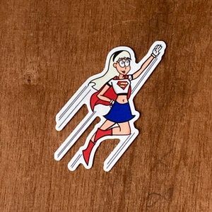 Puede incluir: Una ilustración de dibujos animados de una superheroína que lleva una capa roja, una camiseta blanca con el logotipo de Superman, una falda azul y botas rojas. Está volando con los brazos extendidos.