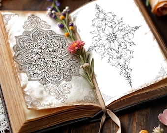 May include: An open book with aged pages featuring a detailed black ink mandala illustration on the left page and a vertical floral scrollwork design on the right page, accented by small pink and yellow flowers placed between the pages.