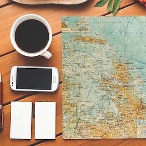 Peut inclure: Une tasse &agrave; caf&eacute; blanche remplie de caf&eacute; noir repose sur une table en bois &agrave; c&ocirc;t&eacute; d'un smartphone blanc, d'un stylo marron, de deux cartes blanches vierges et d'une carte vintage d'une r&eacute;gion c&ocirc;ti&egrave;re.