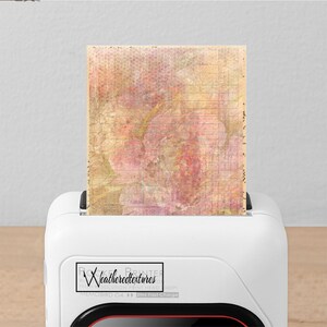 Op de afbeelding: Een witte printer met een rode accent aan de onderkant. De printer heeft een label met de tekst "Rocket Printer by Weatheredtextures High Resolution Print Head 1200x1200dpi Memobird G4 2hrs Fast Charge". Een vel papier met een bloemenontwerp wordt in de printer gevoerd.