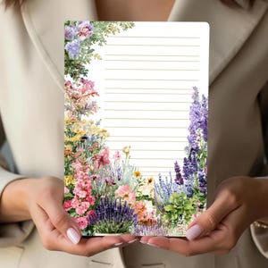 May include: A lined notepad with a floral border featuring pink, purple, yellow, and green flowers. The notepad is held by a person wearing a beige blazer.