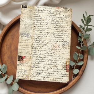 Pu&ograve; includere: Un diario rettangolare con un design a script vintage, appoggiato su un vassoio rotondo di legno. La copertina presenta toni beige e marrone sbiaditi con testo manoscritto e timbri decorativi. Rami di eucalipto verde aggiungono un tocco naturale.