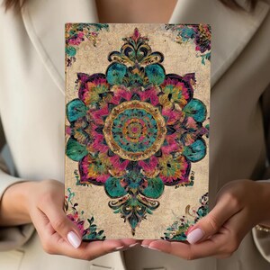 K&ouml;nnte beinhalten: Ein rechteckiges Notizbuch mit einem detaillierten Mandala-Design. Das Mandala weist eine lebendige Farbpalette auf, darunter T&uuml;rkis, Rosa und Gold, vor einem beigefarbenen Hintergrund. Florale Akzente schm&uuml;cken die Ecken. Das Notizbuch wird von einer Person gehalten.