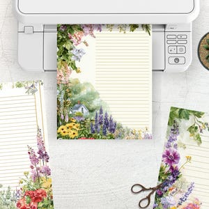 May include: Stationery set with floral designs. The set includes lined paper with a watercolor illustration of a cottage and garden, and additional sheets with floral borders. A printer and a succulent plant are also visible.