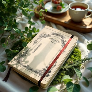 Op de afbeelding: Een crèmekleurig menuboek met een zwart bloemrijk randontwerp en het woord MENU twee keer in het midden gedrukt. Het boek bevat een donkerrood lint als bladwijzer en een bijpassende rode elastische sluiting, rustend op een bed van groen mos.