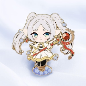May include: A gold-toned enamel pin featuring a stylised character with long white hair, pointed ears, and green eyes. The character wears a white dress with gold trim and holds a staff topped with a red orb. Small blue flowers surround the figure.
