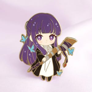 May include: A purple-haired anime-style character pin with a black coat, white dress, and a brown staff. The pin features gold trim and blue butterfly accents. The character has large purple eyes and is holding the staff. The background is a light purple.