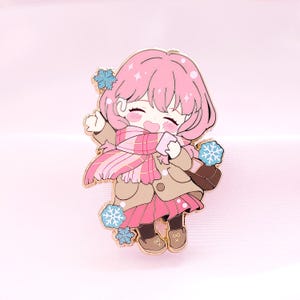 以下が含まれることがあります： 冬の装いの、アニメスタイルの女の子を描いたピンクのエナメルピン。コート、スカーフ、ドレスを着て、カップを持ち、雪の結晶が周りにあります。ピンは金の輪郭と茶色のサッチェルが付いています。
