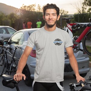 Puede incluir: Un hombre con una camiseta gris con la palabra "excursion" impresa en ella, se encuentra frente a un coche plateado con dos bicicletas sujetas a la parte trasera. El hombre sonríe y mira a la cámara.
