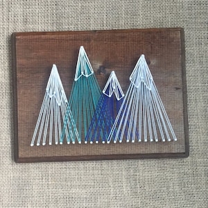 May include: Wooden wall art featuring a mountain range design. The mountains are crafted with white, teal, and blue string, creating a textured effect against the dark brown wood. The artwork is rectangular and suitable for home decor.
