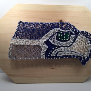 May include: A string art piece depicting the Seattle Seahawks logo. The logo is made of blue, white, and green string wrapped around nails hammered into a wooden board.