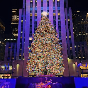 Può includere: Un grande albero di Natale illuminato si erge di fronte a un edificio alto con un bagliore viola. L'albero è decorato con luci colorate e ornamenti. Una statua dorata di una figura che pattina si trova in primo piano.