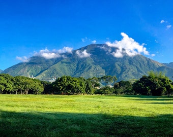 May include: A lush green field with a mountain range in the background. The mountain range is covered in clouds and the sky is a bright blue.