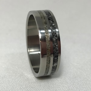 Pode incluir: Um anel de titânio prateado com duas bandas incrustadas de material salpicado de cinza e branco.