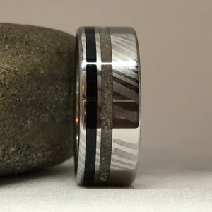 Pode incluir: Uma aliança de casamento prateada e preta com uma linha preta fina e uma faixa de material cinza claro.