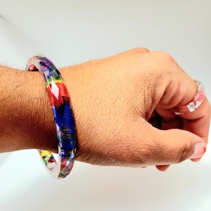 Könnte beinhalten: Ein farbenfrohes Armreif-Armband aus Harz und ein passender Ring. Das Armband zeigt eine lebendige Mischung aus roten, blauen, gelben und violetten Farbtönen. Der Ring hat ein rosa und klares Design. Der Schmuck wird an einer Hand getragen.