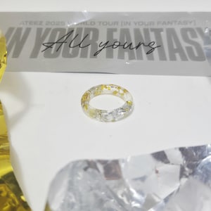 Puede incluir: Un anillo de resina transparente con escamas doradas y plateadas. El anillo es circular y está sobre una superficie blanca. El texto de fondo dice "ATEEZ 2025 WORLD TOUR [IN YOUR FANTASY]" y "All yours."