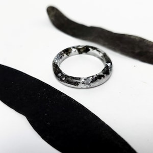 May include: A clear resin ring with embedded silver foil and black accents. The ring has a rounded shape and a smooth, polished finish. The ring is set against a white background with black and gray abstract shapes.