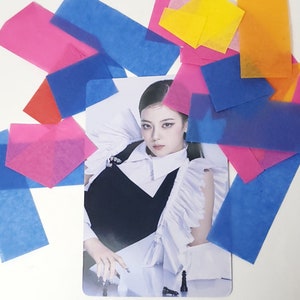 May include: A photo card featuring a woman with dark hair and makeup, wearing a black and white outfit with a ruffled collar. She is sitting at a table with chess pieces on it. The photo card is surrounded by colorful tissue paper.