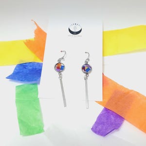 Può includere: Un paio di orecchini pendenti in argento con cabochon di vetro con motivi geometrici colorati. Gli orecchini sono su uno sfondo bianco con carta velina colorata.
