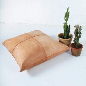 May include: A large, square, tan leather floor cushion with stitched detailing. Two potted cacti are to the right of the cushion. The cushion is a neutral color and would complement many interior design styles.