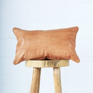May include: A rectangular, light brown leather pillow with rounded corners rests on a three-legged wooden stool. The pillow has a natural, textured appearance, and the stool is made of light-colored wood.