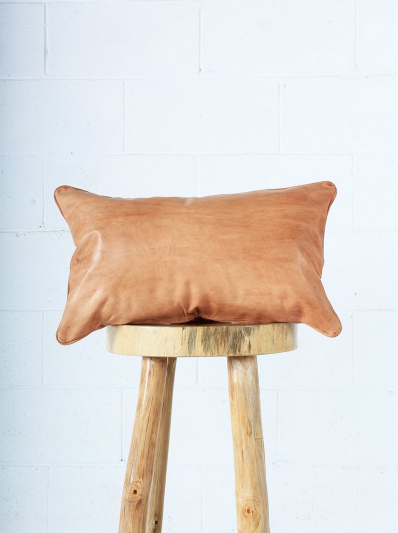 May include: A rectangular, light brown leather pillow with rounded corners rests on a three-legged wooden stool. The pillow has a natural, textured appearance, and the stool is made of light-colored wood.
