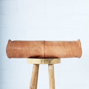 May include: A cylindrical, light brown leather bolster pillow with rounded ends, resting on a three-legged wooden stool. The pillow has a seam running down the center. The stool is made of light-colored wood.