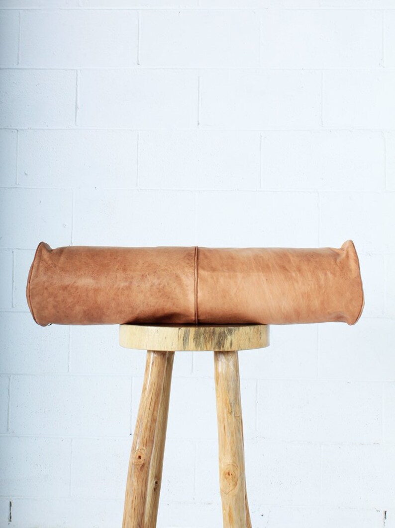 May include: A cylindrical, light brown leather bolster pillow with rounded ends, resting on a three-legged wooden stool. The pillow has a seam running down the center. The stool is made of light-colored wood.