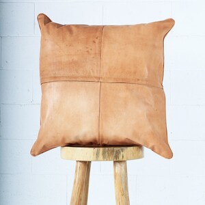 May include: A square, tan leather pillow with a stitched design, resting on a small, light-colored wooden stool. The pillow has a patchwork appearance, with visible seams. The stool has three legs and a round top.