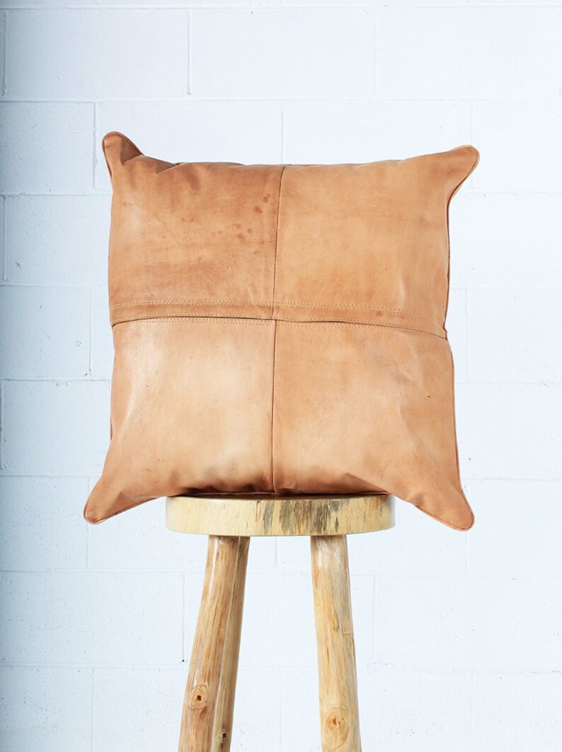 May include: A square, tan leather pillow with a stitched design, resting on a small, light-colored wooden stool. The pillow has a patchwork appearance, with visible seams. The stool has three legs and a round top.