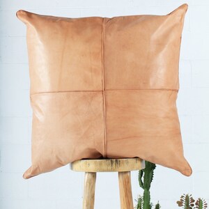 May include: A square, tan leather pillow with a stitched cross pattern, resting on a wooden stool. The pillow has a soft, textured appearance and is a neutral color. The background is a white brick wall.