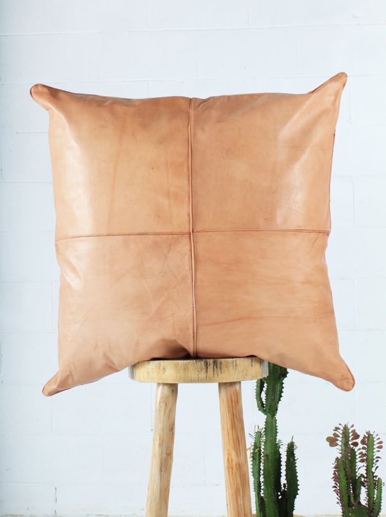 May include: A square, tan leather pillow with a stitched cross pattern, resting on a wooden stool. The pillow has a soft, textured appearance and is a neutral color. The background is a white brick wall.