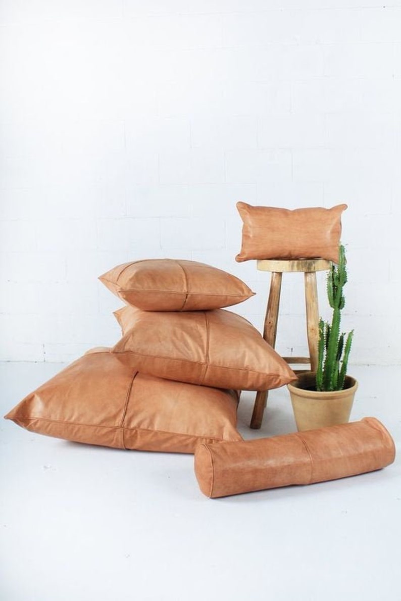 May include: A collection of tan leather pillows in various shapes and sizes. There are several square pillows, a bolster pillow, and a small rectangular pillow on a wooden stool. A potted cactus is also present.