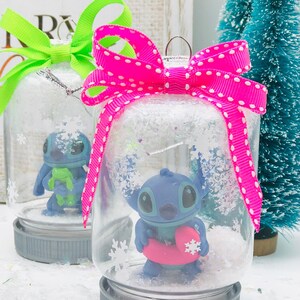 May include: Two glass snow globes with gray lids and pink and green bows. The snow globes contain a blue Stitch character from Lilo & Stitch. One Stitch is holding a green frog and the other is holding a pink inner tube.