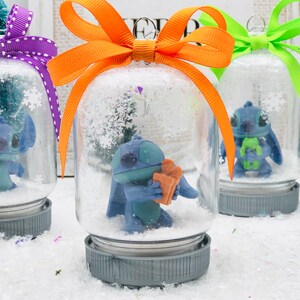 May include: Three glass snow globes with blue Stitch characters inside. The snow globes have different colored bows: purple, orange, and green. The snow globes are sitting on a white surface.