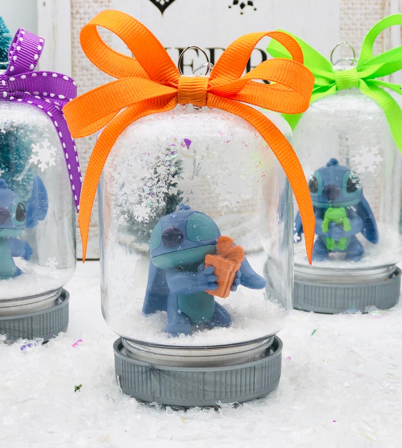 May include: Three glass snow globes with blue Stitch characters inside. The snow globes have different colored bows: purple, orange, and green. The snow globes are sitting on a white surface.
