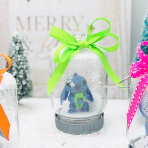 May include: Three glass snow globes with a blue character inside. The snow globes are decorated with green, pink, and white ribbon bows. The snow globes are sitting on a white surface.