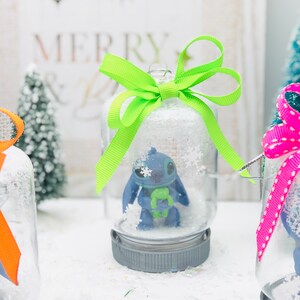 May include: Three glass snow globes with a blue character inside. The snow globes are decorated with green, pink, and white ribbon bows. The snow globes are sitting on a white surface.