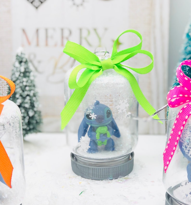 May include: Three glass snow globes with a blue character inside. The snow globes are decorated with green, pink, and white ribbon bows. The snow globes are sitting on a white surface.